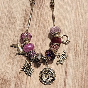 May include: A silver charm bracelet with purple, pink, and red beads. The charms include a dog, a flag, and a round charm with the words "United States Marine Corps". The bracelet also has a charm that says "Marine Wife".