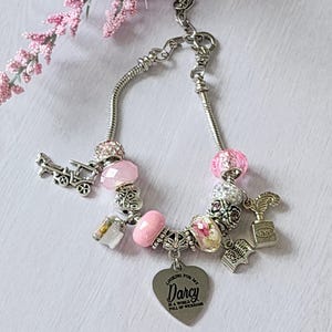 May include: Silver charm bracelet with pink and clear beads, and various charms. Charms include a carriage, a heart with text, a book, and a quill. The bracelet is displayed on a white surface with pink flowers.
