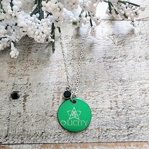 May include: A silver chain necklace with a green round pendant. The pendant has a white star with a pair of glasses inside and the word "OLICITY" written below it. A small black bead hangs above the pendant.