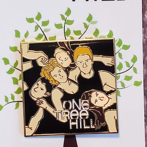 May include: A gold-colored enamel pin featuring a black and white illustration of five people with the text "One Tree Hill" in white lettering.