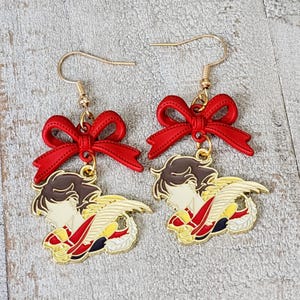 May include: A pair of gold earrings with red bows and a gold charm of a winged figure. The figure is in a profile pose with a red and gold design.