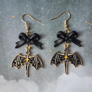 May include: A pair of gold-colored drop earrings. Each earring features a black bow above a gold-colored dragon pendant with black and gold detailing. The dragon pendant has a yellow star in the center.