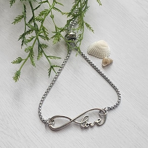 May include: Silver-toned infinity bracelet with the word "love" scripted within the infinity symbol. The bracelet is on a chain with an adjustable clasp. The background includes greenery and seashells, suggesting a beach theme.