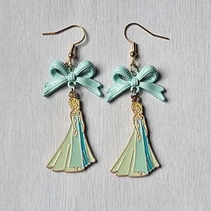 May include: A pair of dangle earrings featuring a gold-tone hook, a light blue enamel bow, and a gold-tone charm depicting a figure with long blonde hair wearing a flowing light blue and mint green gown that reaches the floor.