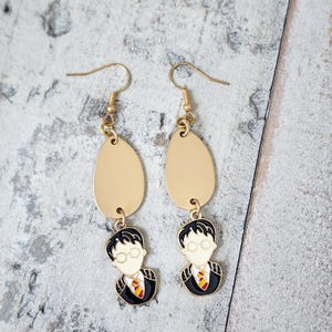 May include: Gold dangle earrings with a black and white image of a boy wearing glasses and a tie. The earrings have a gold teardrop shape.