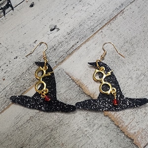May include: A pair of black glitter witch hat earrings with gold glasses charms and red crystal accents.