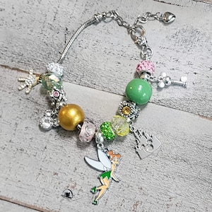 Könnte beinhalten: Ein silbernes Charm-Armband mit grünen, goldenen und rosafarbenen Perlen. Die Charms beinhalten einen Tinkerbell-Charakter, ein Schloss, einen Schlüssel und Mickey-Maus-Ohren.