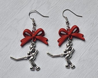 May include: A pair of dangle earrings featuring a silver-toned hockey player charm suspended from a bright red bow. Each earring includes a standard silver-toned fishhook wire for attachment, showcasing a detailed athletic figure in motion.