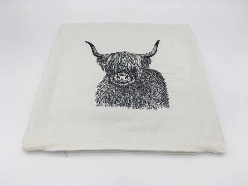 May include: A white canvas pillow with a black and white line drawing of a Highland cow with long horns.