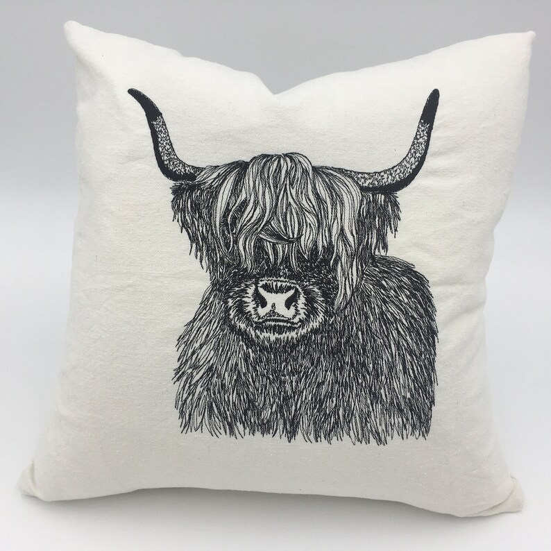 May include: A white throw pillow with a black and white line drawing of a Highland cow with long, shaggy hair. The cow is looking directly at the viewer.