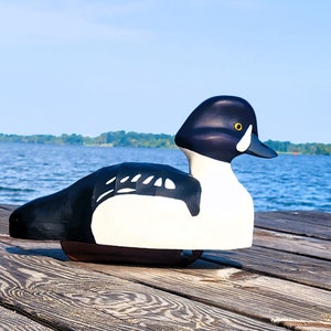 Puede incluir: Un se&ntilde;uelo de pato de madera negro y blanco con un ojo amarillo. El se&ntilde;uelo est&aacute; sentado en un muelle de madera con un lago azul en el fondo.