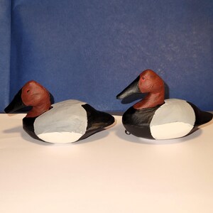May include: Two hand-painted wooden duck decoys, one facing left and one facing right. Both decoys are painted in shades of black, white, gray, and red, with a realistic depiction of a duck's head and body.