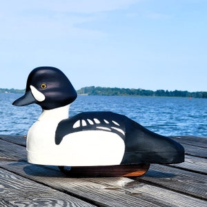 Puede incluir: Un se&ntilde;uelo de pato de madera negro y blanco con un ojo amarillo. El se&ntilde;uelo est&aacute; sentado sobre una superficie de madera.