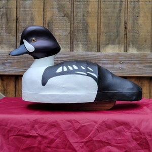 Puede incluir: Un se&ntilde;uelo de madera pintado a mano de un pato goldeneye negro y blanco. El se&ntilde;uelo est&aacute; pintado con detalles realistas, incluyendo un ojo amarillo y marcas negras y blancas en la cabeza y el cuerpo.