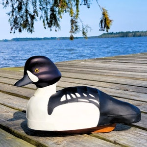 Puede incluir: Un se&ntilde;uelo de pato de madera negro y blanco con un ojo amarillo. El se&ntilde;uelo est&aacute; sentado en un muelle de madera con un lago en el fondo.