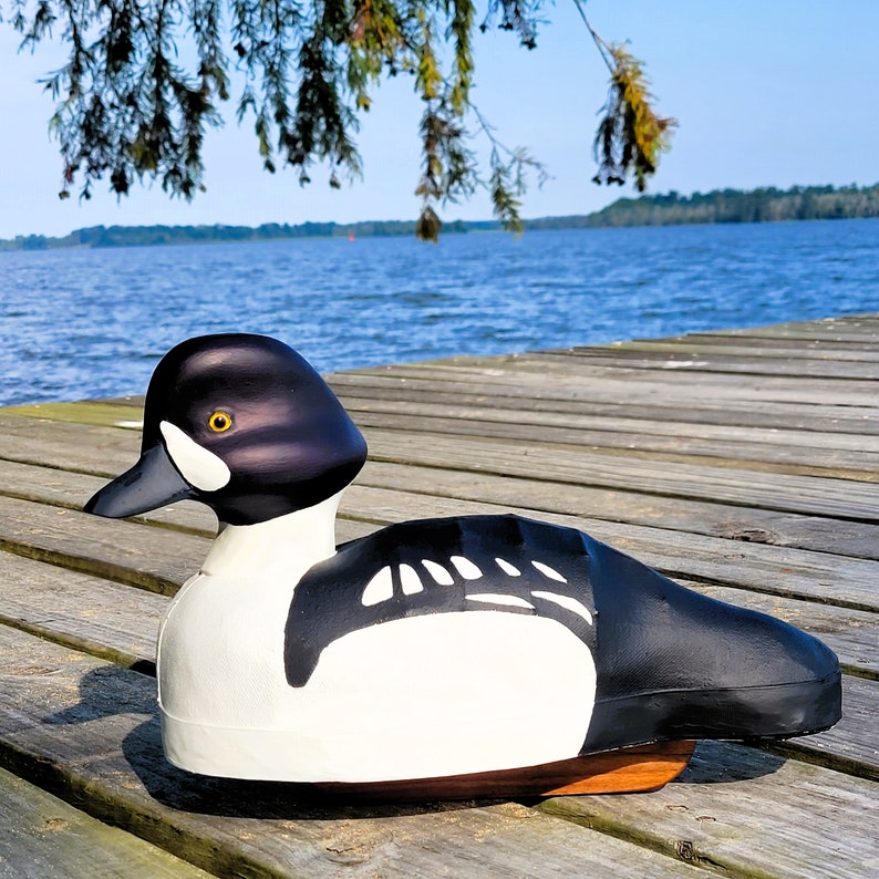 Puede incluir: Un se&ntilde;uelo de pato de madera negro y blanco con un ojo amarillo. El se&ntilde;uelo est&aacute; sentado en un muelle de madera con un lago en el fondo.
