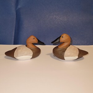 May include: Two wooden duck decoys painted in brown, white, and black. The decoys are facing each other and are positioned on a white surface.