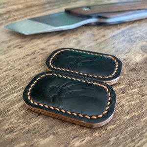 May include: Two black leather keychains with a stitched border and a decorative design. The keychains are on a wooden surface.