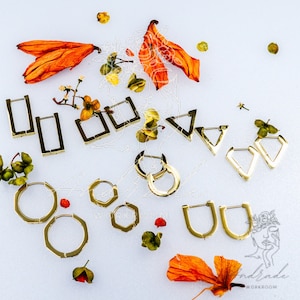 May include: A collection of gold-toned earrings in various shapes, including hoops, squares, and triangles. The earrings are arranged on a white surface with dried orange flowers and leaves.