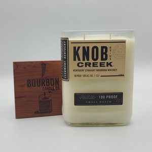 May include: A white candle in a clear glass bottle with a brown label that reads "Knob Creek Kentucky Straight Bourbon Whiskey 100 Proof | 50% Alc/Vol" and "Crafted for Superior Taste & Smoothness 100 Proof Small Batch". The bottle is sitting on a wooden square with the words "Bourbon Candle Co." printed on it.