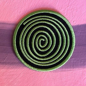 May include: A round, black and green embroidered patch with a spiral design.