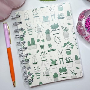 Peut inclure: Un carnet à spirales avec une couverture crème ornée d'un motif de plantes vertes. Un stylo orange et rose est posé à côté du carnet. Autres éléments : une théière, un petit bol et un récipient rose avec des trombones roses.