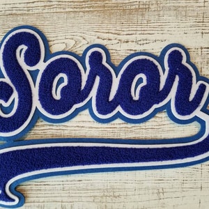 May include: A blue and white felt applique with the word "Sarah" in a cursive font. The applique is shaped like a baseball cap.