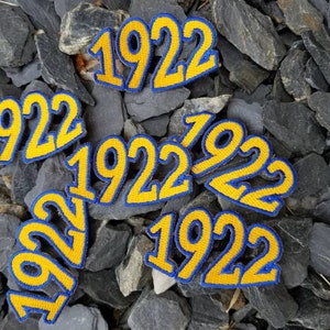 Könnte beinhalten: Mehrere gestickte Patches in Gelb und Blau, auf denen jeweils das Jahr 1922 zu sehen ist. Die Patches sind auf einem Hintergrund aus grauen Steinen angeordnet.