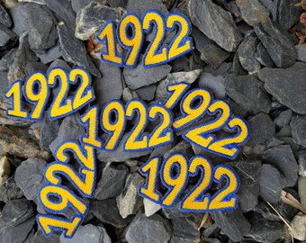May include: Several yellow and blue embroidered patches, each featuring the year 1922. The patches are displayed against a backdrop of grey stones.