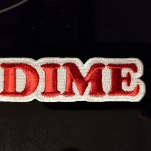 Puede incluir: Parche bordado rojo con la palabra "DIME" en letras blancas con contorno.