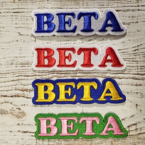 Puede incluir: Cuatro parches bordados con la palabra "BETA" en diferentes colores. Los parches son blancos con letras azules, rojas, amarillas y rosas. Los parches están sobre un fondo de madera clara.
