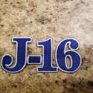 May include: Blue and white embroidered patch with the text "J-16" in a retro style font.
