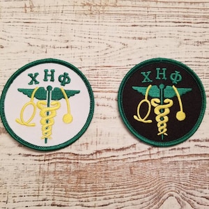 May include: Two round embroidered patches with green borders. The patches feature a caduceus medical symbol with a snake and wings, and the letters XHΦ. One patch has a white background and the other has a black background.