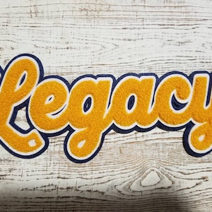 May include: A yellow felt patch with blue outlining, spelling out the word "Legacy" in a cursive font.