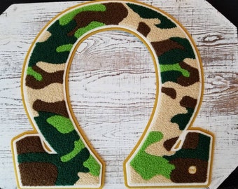 May include: A large, green, brown, and beige felt letter "O" with a yellow outline. The letter is in a camouflage pattern.