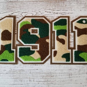 May include: A green, brown and white camouflage fabric patch with the number "1911" in white outlined in brown. The patch is made of a fuzzy material and has a white border.