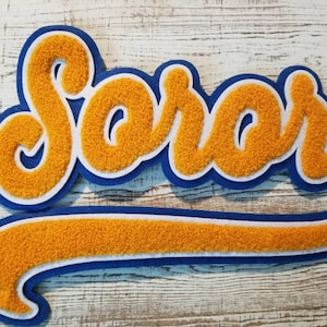 Op de afbeelding: Een blauwe en witte vilt applicatie met het woord "SOROR" in gele vilt letters. De applicatie is gevormd als een baseball team logo.