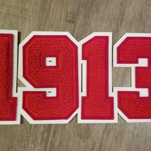 May include: Red felt applique numbers spelling out "1913" with a white border. The numbers have a textured, fuzzy appearance. The "3" has a small registered trademark symbol.