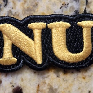 Puede incluir: Parche bordado con las letras "NU" en hilo dorado sobre fondo negro. El parche tiene un borde negro. Las letras están en una fuente audaz y estilizada. Diseñado para aplicar en ropa o accesorios.