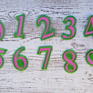 May include: A set of embroidered number patches, displaying digits 0 to 9. Each number has a green interior with a pink border. They are arranged on a white, distressed wooden surface. Ideal for crafting or customisation projects.