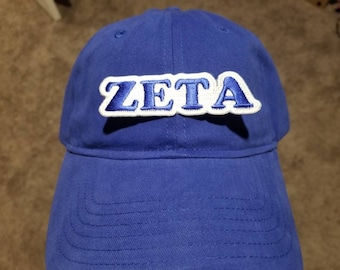 Peut inclure: Une casquette de baseball bleue avec un patch brodé blanc et bleu qui dit "ZETA".