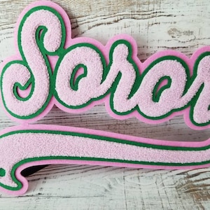 May include: Pink and green felt applique with the word "Soror" in a cursive font.