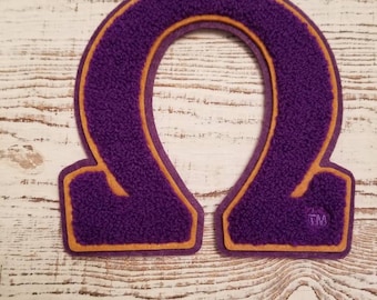 May include: Purple felt letter Omega with a gold outline. The letter is on a white wooden background. The letter has a trademark symbol in the bottom right corner.