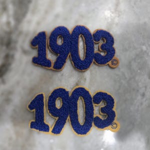 Peut inclure: Deux écussons brodés avec le numéro "1903" dans une police audacieuse, bleue et texturée. Les chiffres sont soulignés en couleur beige. Les écussons sont sur une surface de marbre gris clair et blanc.