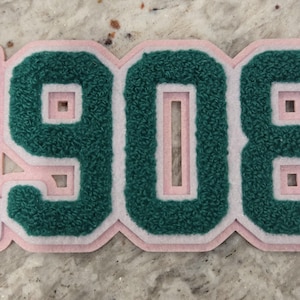 以下が含まれることがあります： ピンクと白のフェルトパッチに緑色の数字1908が書かれています