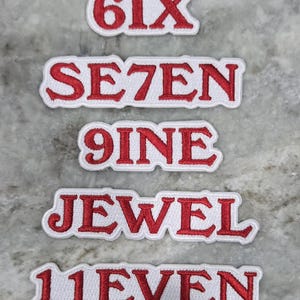 May include: Embroidered patches with red text on a white background. The patches display the words "FIVE", "SIX", "SE7EN", "9INE", "JEWEL", "11EVEN", and "12ELVE". The patches are likely for clothing or accessories.