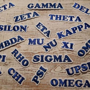 May include: A collection of blue and white embroidered patches with the Greek alphabet letters. The patches are arranged on a light wood surface.