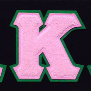 May include: Pink letters spelling "AKA" with a green outline. The letters are raised and have a fuzzy texture.