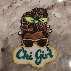 May include: An embroidered patch featuring a cartoon illustration of a Black woman with a bun, sunglasses, and a yellow bow. The text "Chi Girl" is written in green cursive on a white background with a yellow outline.