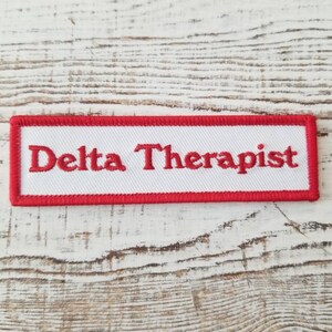 May include: A rectangular white patch with the words "Delta Therapist" embroidered in red. The patch has a red border and is set against a light-colored, textured wooden surface.